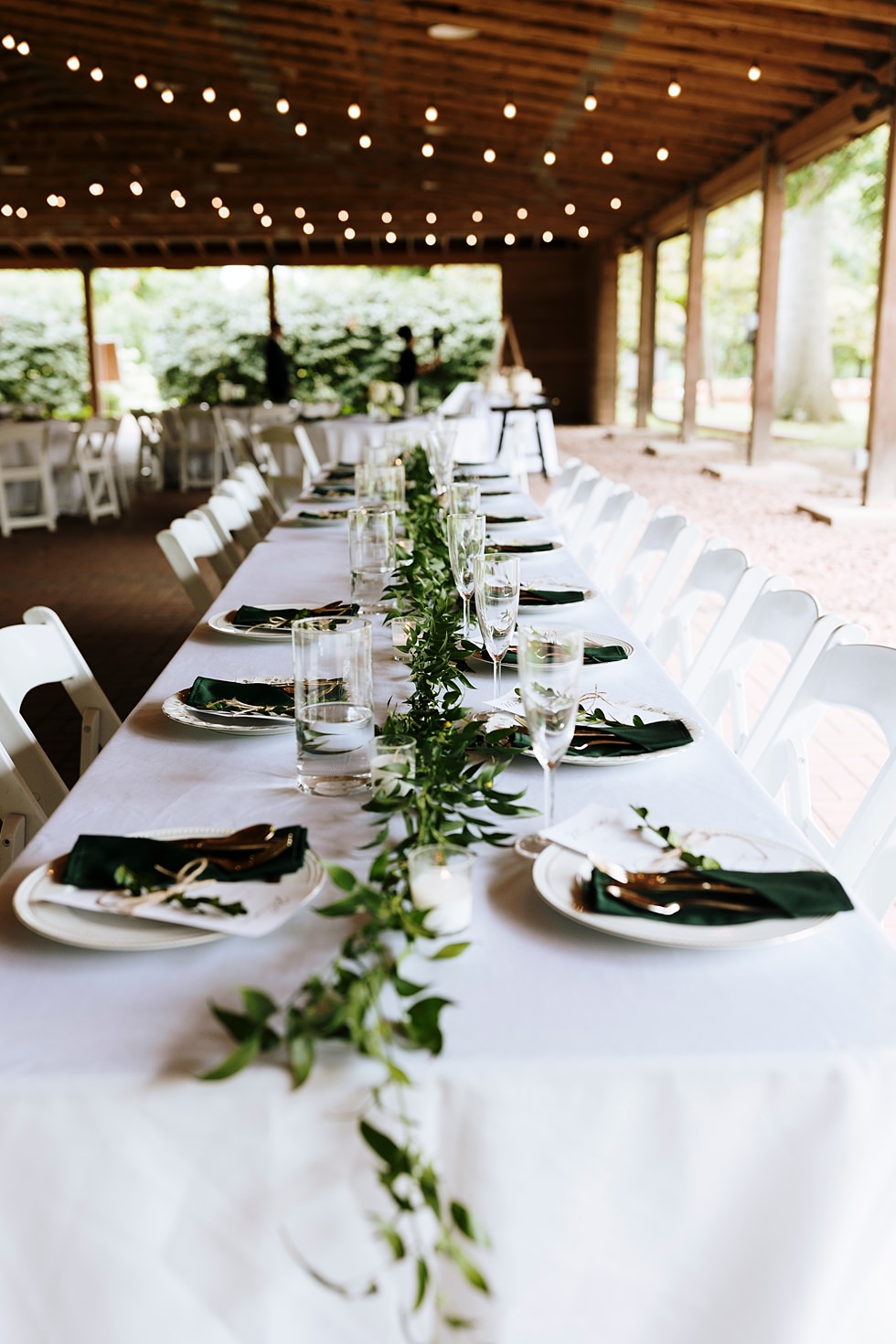 Long wedding reception table decorated with greenery, candles, and dark green napkins.