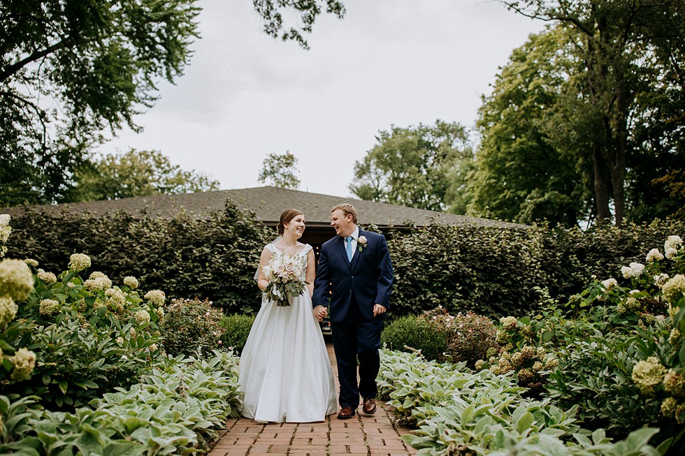 Bride and groom walk hand in hand through a lush hydrangea garden at Farmington Historic Home wedding venue.