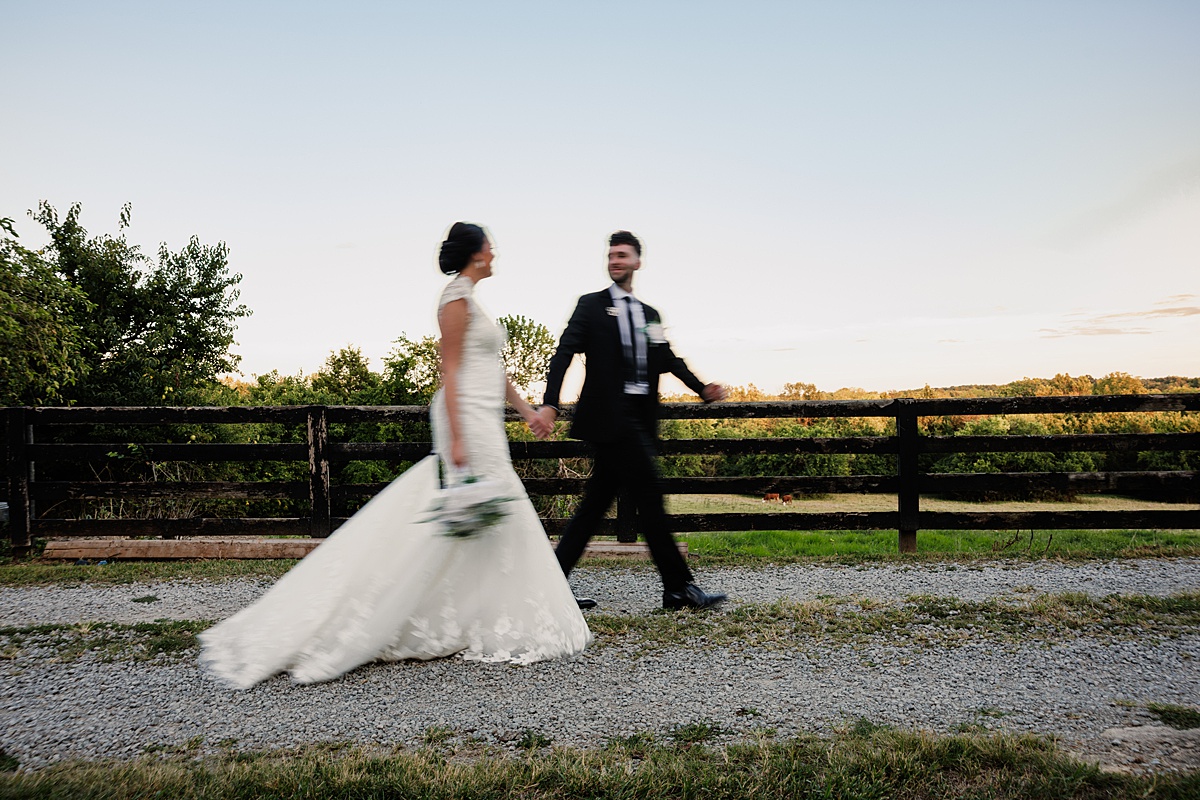 Bride and groom walking hand in hand beside a fenced pasture at sunset.