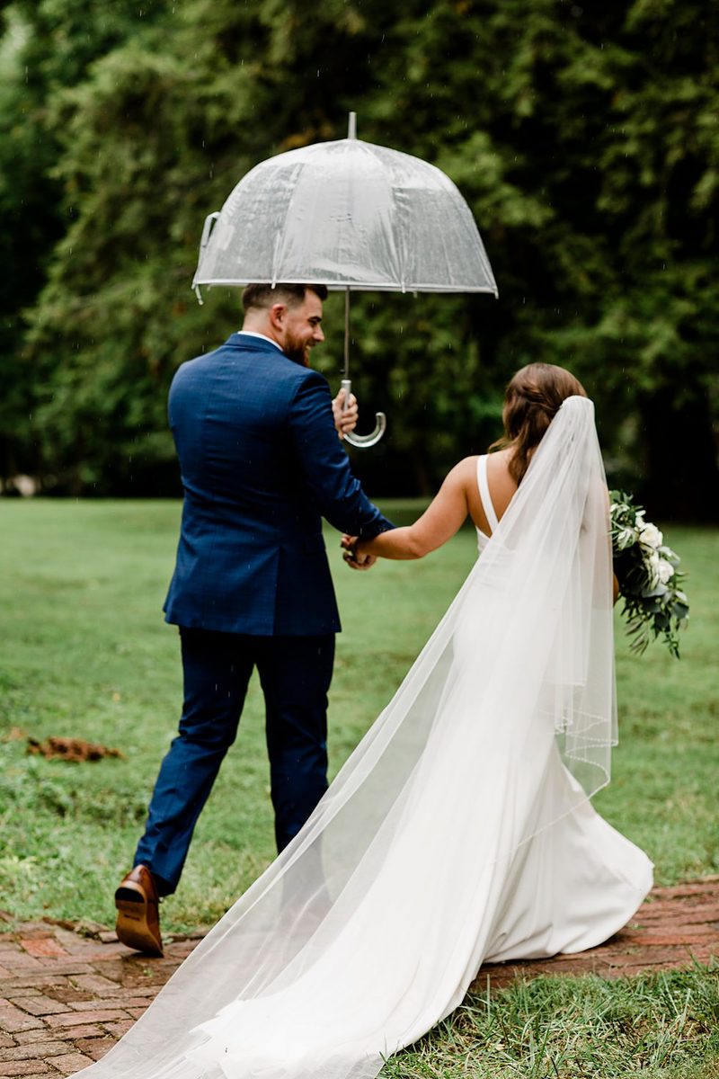 Bride and groom walk hand in hand beneath a clear umbrella on their rainy wedding day.