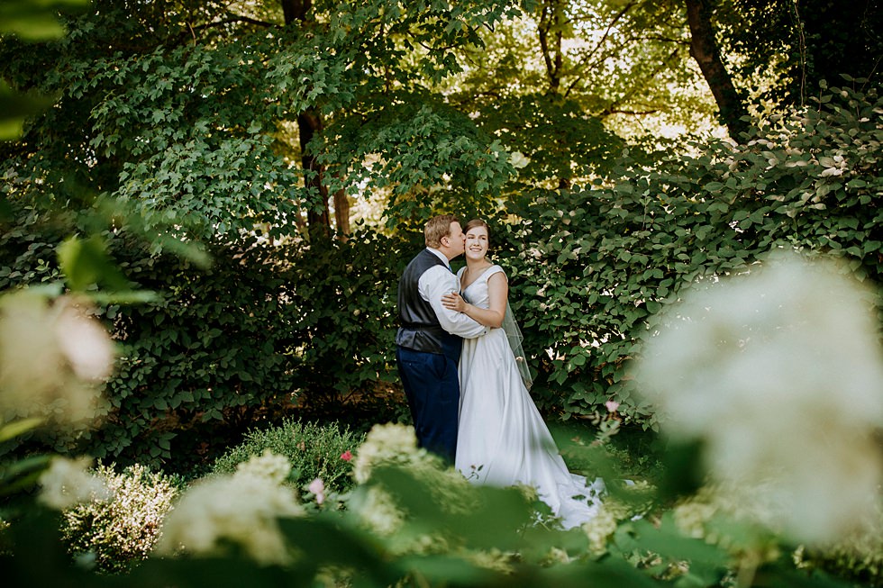 Groom kisses the bride’s cheek during a garden wedding portrait surrounded by greenery.
